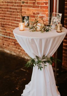 a white table topped with pictures and greenery next to a brick wall in front of a window