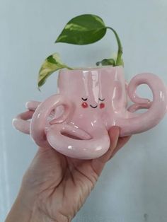 a hand holding a pink ceramic cup shaped like an octopus with eyes closed and a plant in it's mouth