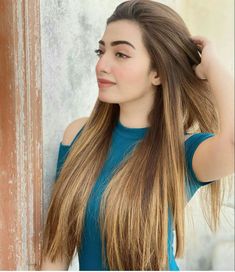 a woman with long hair standing next to a wall wearing a blue shirt and holding her hand on her head