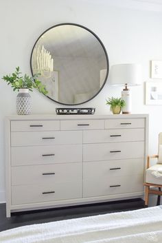 a white dresser with a round mirror above it and a plant on the top shelf