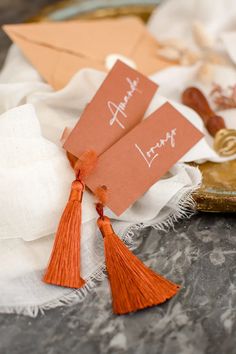 two orange tasseled tags on top of a white table cloth next to an envelope