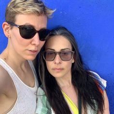 two women standing next to each other in front of a blue wall with sunglasses on