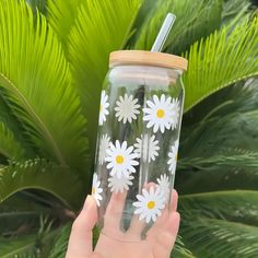 a hand holding a glass with white daisies on it and a straw in the cup