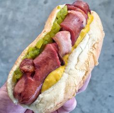 a hand holding a hot dog with bacon and mustard on it's buns