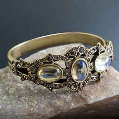 "This rich, unusually impressive bracelet is created from 90 year old stones set into our studio's original sculpture. The bracket is finely detailed and set with premium Swarovski smoke crystal. Three oval stones are vintage beauties from the 1930s which I acquired from a former exporter in Czechoslovakia. The bracket is mounted on a split shank cuff and will comfortably fit 7 to 8\" wrists. Hand burnished warm bronze finish. Entirely made in our Los Angeles studio. Choices:  AMETHYST EMERALD R Bohemian Gemstone Wedding Bangle, Collectible Gemstone Bangle Bracelets, Antique Adjustable Jeweled Jewelry, Unique Collectible Jeweled Jewelry, Antique Gemstone Bracelet For Anniversary, Unique Brass Wedding Bracelets, Antique Adjustable Bracelets With Jewels, Unique Brass Bracelets For Wedding, Antique Jeweled Bangle