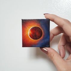 a hand holding up a small square piece of art with the image of a planet in it