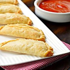 three pastries on a white plate next to a bowl of tomato sauce and a red checkered napkin