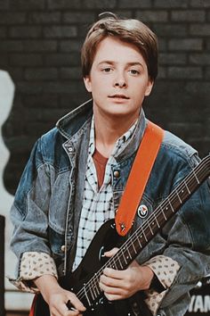 a young man holding an electric guitar in front of a brick wall and wearing a denim jacket