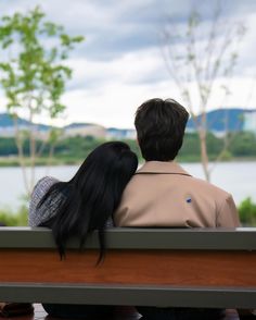 two people are sitting on a bench looking at the water from behind their backs, with trees in the background