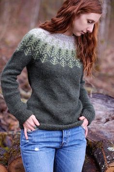 a woman standing next to a pile of logs in the woods wearing a green sweater