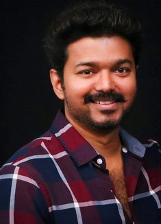 a man with a mustache smiling at the camera in front of a black background and wearing a checkered shirt