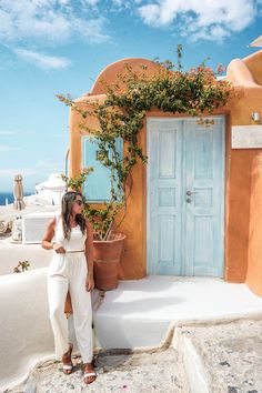 a woman standing in front of a blue door on the side of a building with potted plants