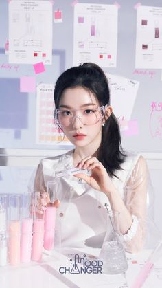 a woman in glasses sitting at a table with pink pens and paper notes on the wall behind her