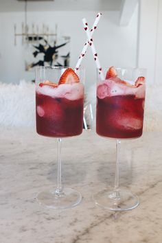 two glasses filled with liquid and strawberries on top of a marble counter next to a clock