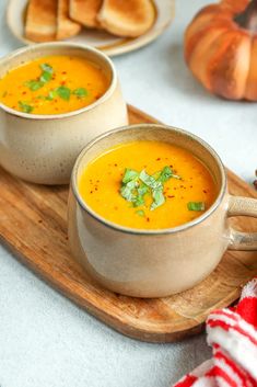 two bowls of soup are sitting on a wooden tray next to some bread and pumpkins