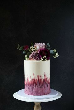 a white and pink cake with purple flowers on top sitting on a stand in front of a black wall