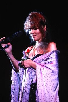 a woman holding a microphone in her right hand and wearing a purple shawl with polka dots on it