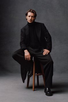 a man sitting on top of a wooden chair wearing a black suit and turtle neck sweater