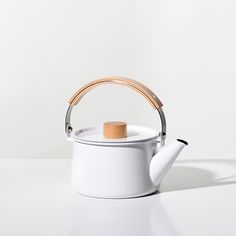 a white teapot with a wooden handle and lid on a plain surface, sitting in front of a wall