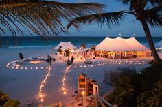 people are gathered on the beach at night for an outdoor wedding reception with white tents and string lights