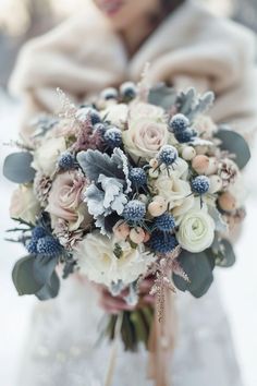 a bridal holding a bouquet of white and blue flowers in the snow with a woman standing behind her