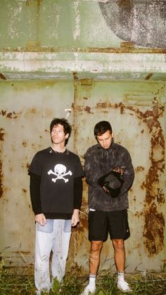 two men standing next to each other in front of a rusted wall with a skull on it