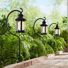 three lights are hanging on the side of a brick walkway in front of trees and bushes