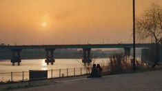 two people sitting on the side of a river watching the sun go down in the distance