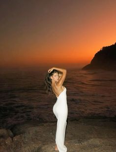 a woman standing on top of a rock next to the ocean at sunset with her arms behind her head