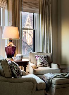 a living room with two chairs and a table in front of a window that has curtains on the windowsill