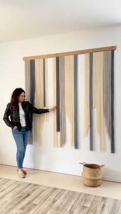 a woman standing next to a wall hanging on the side of a white wall with long strips of yarn