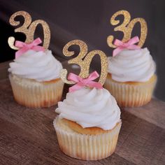 three cupcakes with white frosting and pink bows are on a wooden table