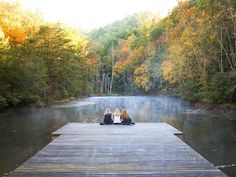 three women sitting on a dock in front of a body of water surrounded by trees