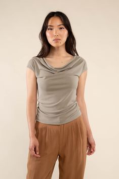 a woman standing in front of a white wall wearing brown pants and a gray top