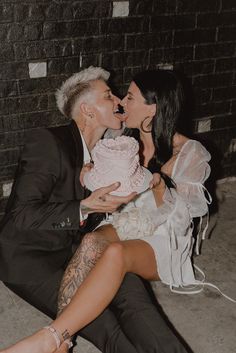 a man and woman sitting on the ground kissing while holding a cake in front of them