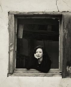 a black and white photo of a woman in a window looking at the camera with her hand on her chin