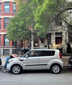 two people sitting on the back of a silver minivan parked in front of a building