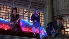 three people on stage playing instruments and singing into microphones in front of a neon sign