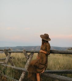 a woman in a brown dress and hat leaning on a wooden fence