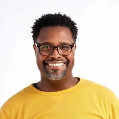 a man with glasses and a yellow shirt smiles at the camera while standing in front of a white background