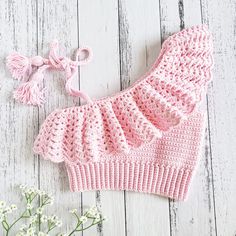 a pink knitted baby outfit next to flowers on a white wooden background with a bow