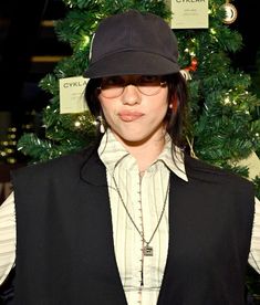 a woman wearing a black hat and vest in front of a christmas tree with tags on it