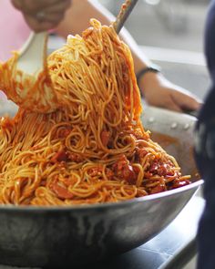 someone is stirring noodles in a pan on top of the stove, while another person uses a spoon to stir them