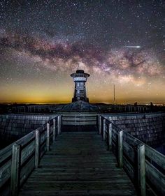 the night sky is filled with stars above a wooden walkway leading to a light house