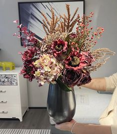 a woman holding a vase with flowers in it on a table next to a dresser