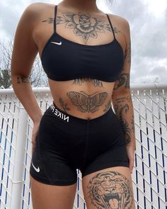 a woman with tattoos standing next to a fence wearing black shorts and a sports bra