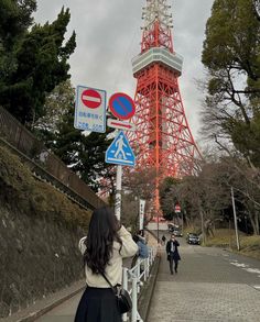 a woman standing next to a street sign near a tall red tower in the distance