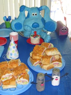 a blue table topped with lots of sandwiches and other food on plates next to a stuffed animal