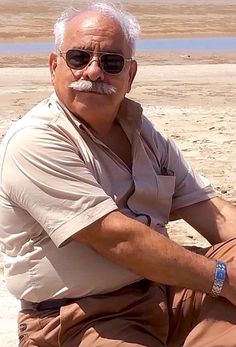 an older man sitting on the beach wearing sunglasses and a moustache with a mustache