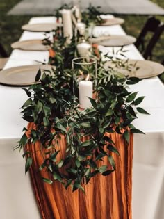 a long table with candles and greenery on it is set for an outdoor dinner